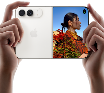 iPhone Duo, สีสตาร์ไวท์, ถืออยู่ในมือสองข้างในแนวนอน, กางเปิดอยู่, แผงด้านซ้ายแสดงกล้องหลัง Dual Fusion 48MP, แผงด้านขวาแสดงบุคคลหนึ่งขณะถ่ายภาพบนจอภาพภายนอก, ตัวแบบในภาพสามารถเห็นใบหน้าของตัวเองระหว่างกำลังถ่ายภาพโดยใช้ Duo Preview