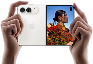iPhone Duo, สีสตาร์ไวท์, ถืออยู่ในมือสองข้างในแนวนอน, กางเปิดอยู่, แผงด้านซ้ายแสดงกล้องหลัง Dual Fusion 48MP, แผงด้านขวาแสดงบุคคลหนึ่งขณะถ่ายภาพบนจอภาพภายนอก, ตัวแบบในภาพสามารถเห็นใบหน้าของตัวเองระหว่างกำลังถ่ายภาพโดยใช้ Duo Preview